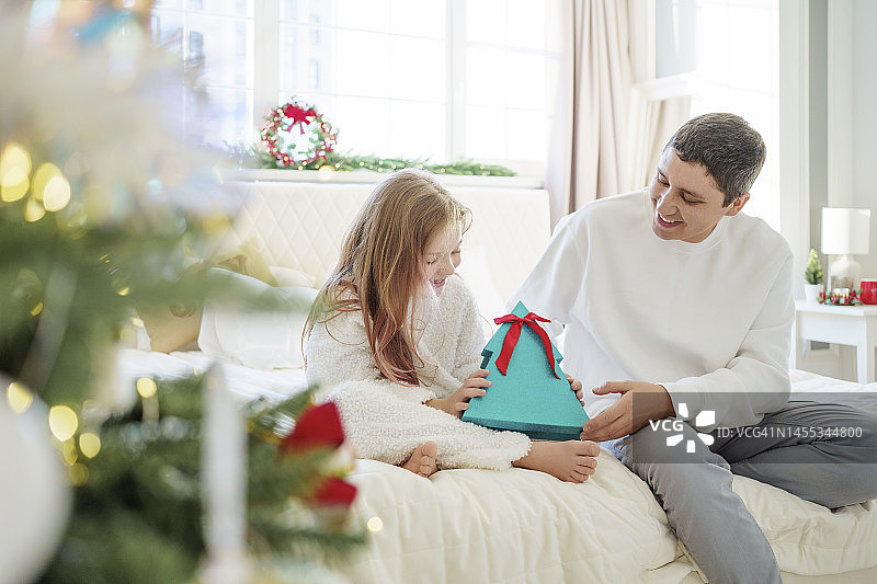 快乐的女孩拿着圣诞礼物和爸爸坐在家里的床上图片素材