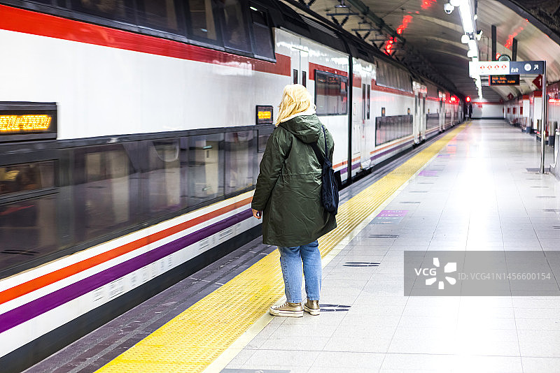 胖乎乎的金发女人，穿着绿色外套，在车站等火车停下来。旅行、火车、交通工具、可持续性、火车站和旅行的概念。图片素材