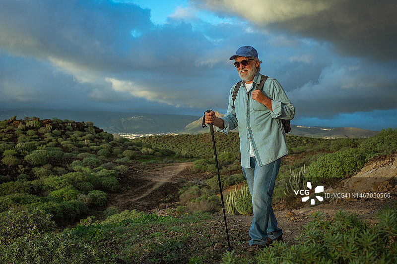 快乐的老活跃的老人享受在夕阳下的乡村徒步旅行。年长的白人男性，戴着帽子，背着背包走在人行道上图片素材