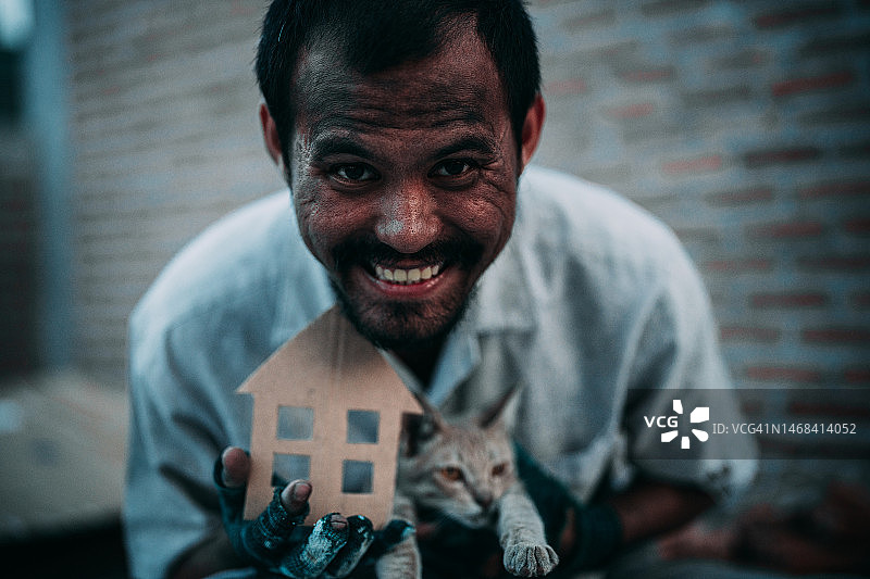 亚洲无家可归的人抱着他的猫在一片荒地上，抱着一个房子形状的纸箱。图片素材