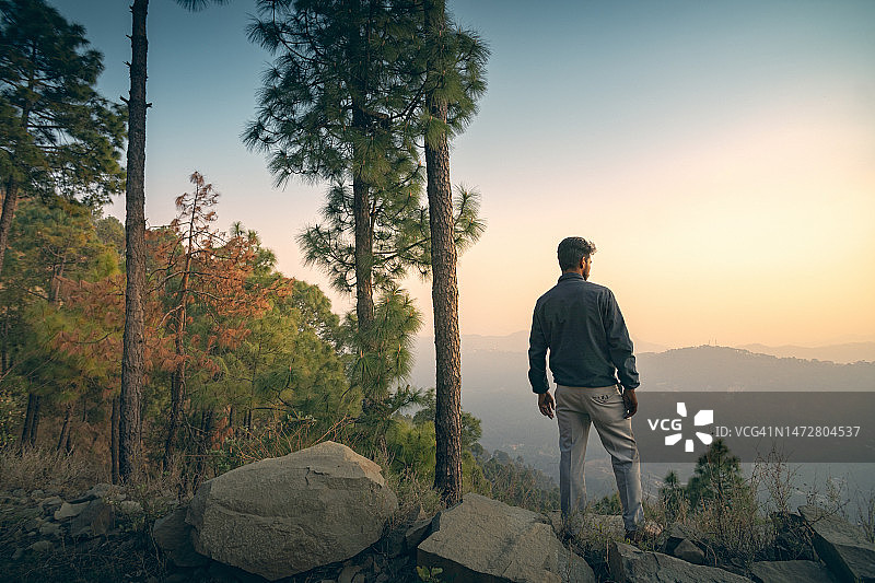 一个年轻人站在山上的一块岩石上看日落。图片素材
