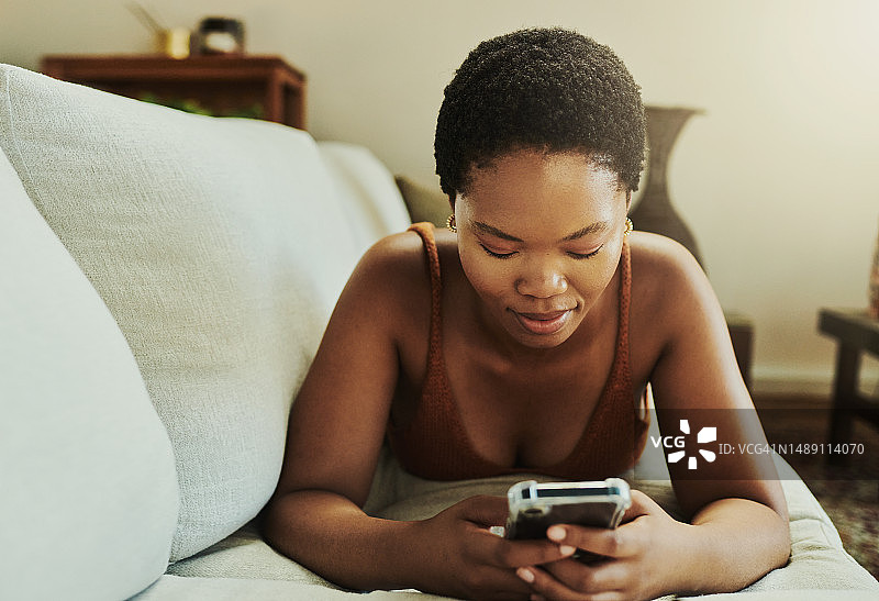 年轻的黑人女性躺在沙发上，一边翻着手机，一边微笑着看照片。图片素材