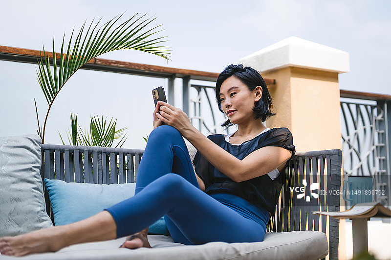 年轻的亚洲女子舒适地坐在躺椅上，在阳台上放松，用智能手机发短信图片素材