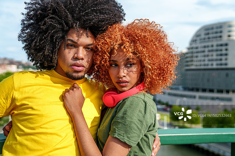 充满活力的拉丁二人组:年轻、时尚、非洲头发的夫妇戴着耳机在一座城市的桥上拍照。图片素材