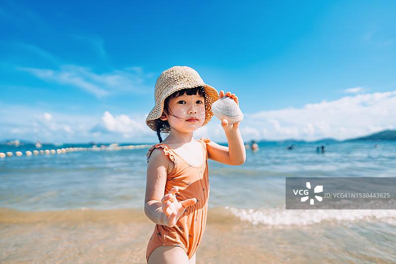 一个可爱的亚洲小女孩戴着遮阳帽，在海滩上拿着一个巨大的蛤蜊壳。微笑着看着镜头。在一个阳光明媚的夏日，在海滩上玩得很开心图片素材