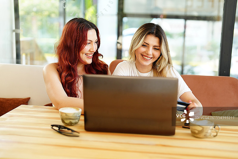 两个快乐的年轻女子坐在咖啡馆的桌子旁，用笔记本电脑上网购物。网上购物概念图片素材