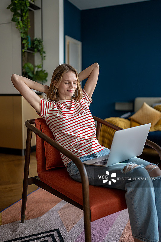 无忧无虑的女孩坐在椅子上，用电脑休息，带着满意的微笑向后靠。图片素材