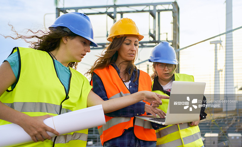 三名多种族女性电力工人的低角度前视图，穿着安全装备和安全帽，在发电厂的背景下，在风力涡轮机过程中看着相机图片素材