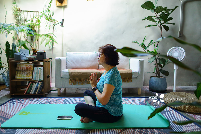 一位年长的亚洲妇女坐在瑜伽垫上，按照手机上的瑜伽指导，冥想，放松，寻找内心的平静。图片素材