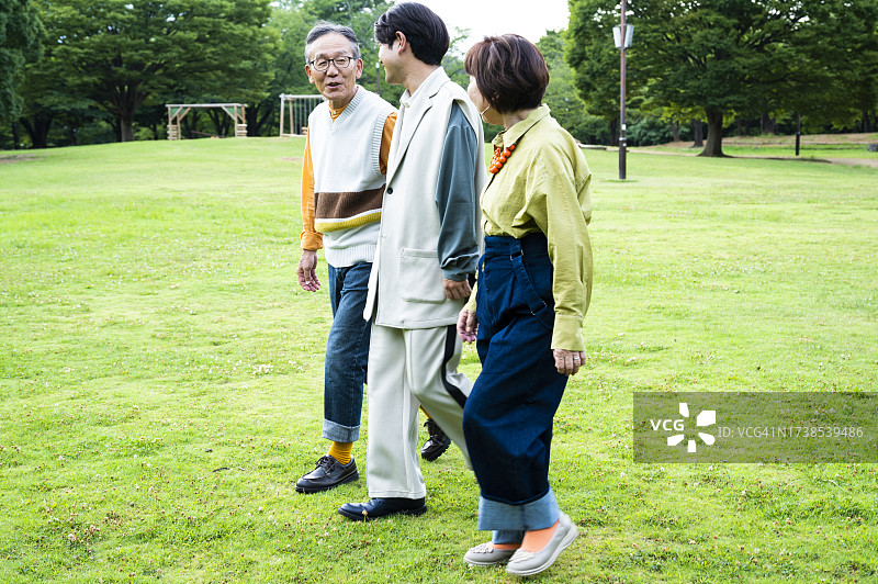 一个成年男子和他年迈的父母在美丽的绿色户外散步，开心地聊天，笑着。丰富的生活。现代家庭场景。图片素材