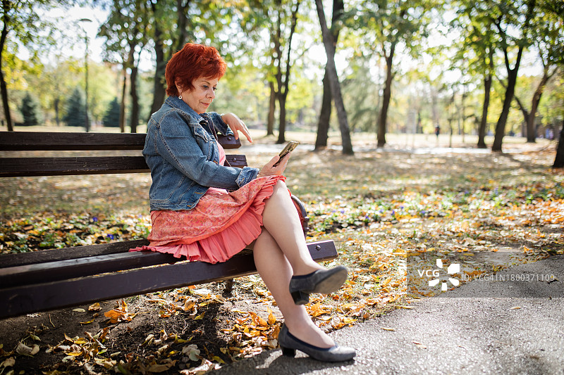 红头发的老女人，坐在公园的长椅上玩她的智能手机。晴朗的秋日。图片素材