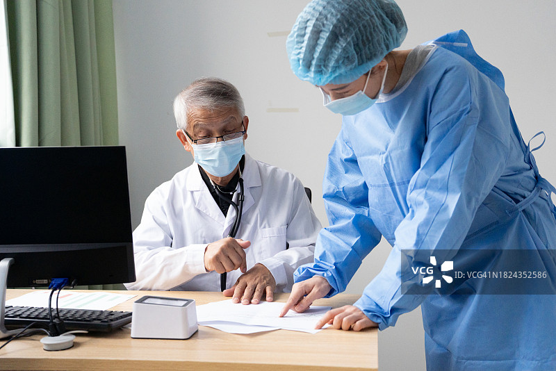 医生和护士在医疗机构的照片。医生坐在桌旁，护士站在他旁边。护士穿着一件蓝色的手术服和手术帽。医生穿着一件白色的实验室大褂。图片素材
