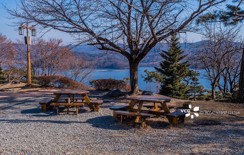 在光秃秃的树前摆着两张圆形的木制野餐桌，背景是湖泊和山脉，韩国，韩国，韩国，亚洲图片素材