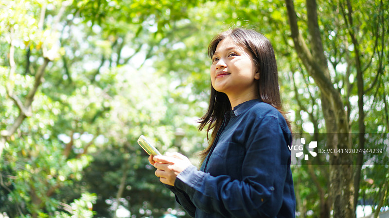 快乐的年轻亚洲女子在公园里，一边用智能手机发短信，一边享受着城市户外阳光明媚的日子图片素材