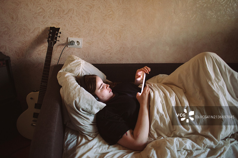 年轻女子戴着耳机躺在床上玩智能手机图片素材