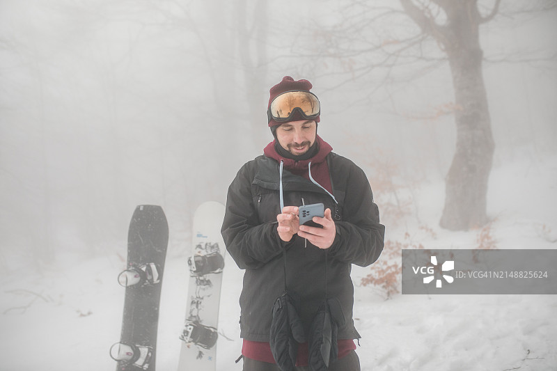 面带微笑的男子穿着滑雪装备，戴着安全眼镜和智能手机。滑雪运动员在山上徒步旅行图片素材