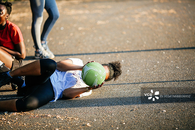 一个年轻的女人在户外的球场上胡闹，侧躺着，拿着一个无篮网球挡在脸上图片素材