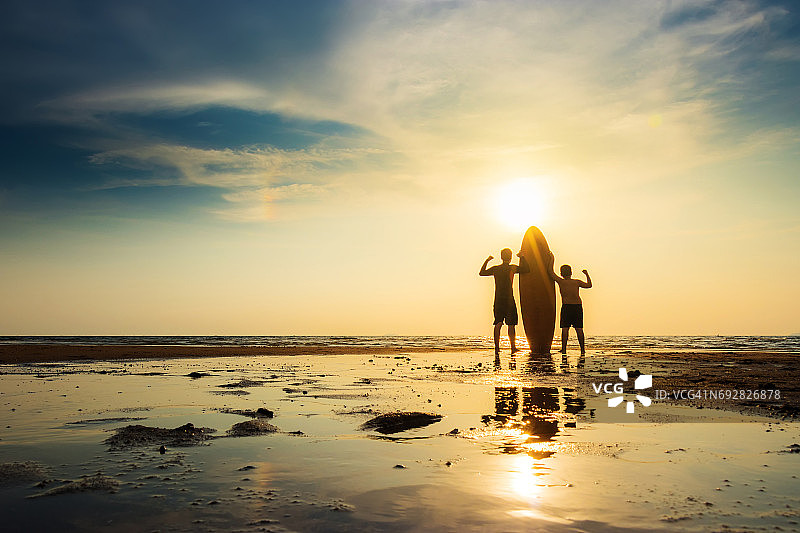 冲浪男子的剪影和一个男孩站着冲浪板，手向上。在日落海滩冲浪。户外水上运动冒险生活方式。夏天的活动。图片素材