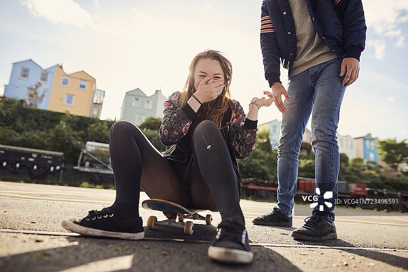 两个朋友在户外闲逛，年轻女子坐在滑板上，笑，低节，布里斯托尔，英国图片素材