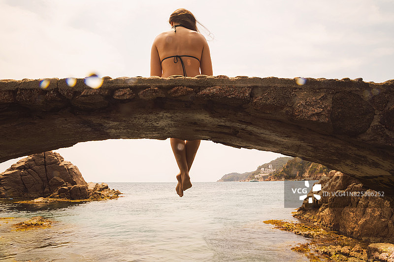 快乐的女人穿着比基尼坐在和凝视科斯塔布拉瓦水与美丽的日落光在夏天在加泰罗尼亚地区旅游度假。图片素材