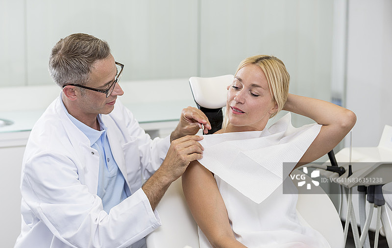牙科美容诊所- - -身穿白大褂的中年牙医在椅子上咨询一头金色长发的女病人，医生拿起餐巾图片素材