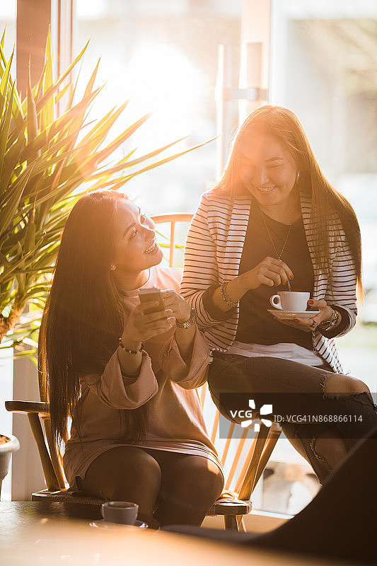 快乐的亚洲女人显示手机短信给她的朋友在咖啡馆。图片素材