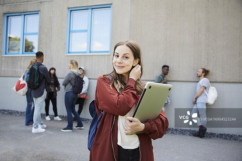 微笑的女学生拿着笔记本电脑站在校园里，背景是朋友图片素材