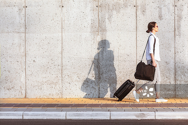 一名年轻女子走在水泥墙边的人行道上，拉着一个带轮子的小行李图片素材