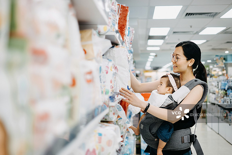 年轻的亚洲母亲抱着可爱的女婴在购物中心购买婴儿产品，正在看各种尿布图片素材