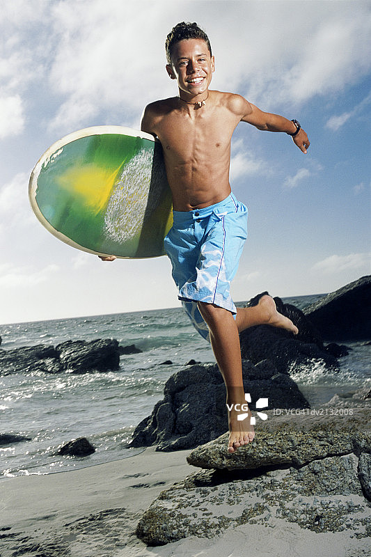 一个无忧无虑的十几岁的男孩沿着海滩跑步的肖像，带着一个冲浪板图片素材