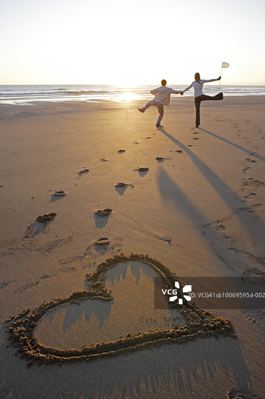 年轻夫妇在沙滩上跳跃，在沙子上画着心形符号图片素材