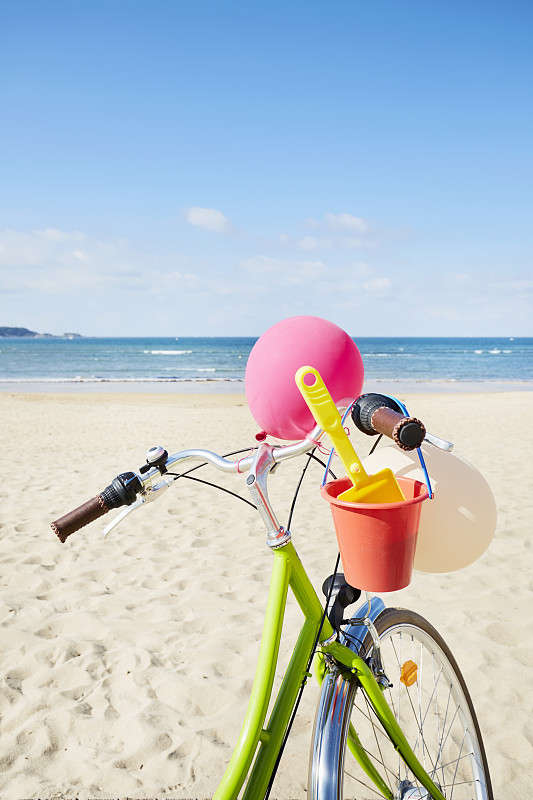 绿色自行车与气球和沙桶和铲子在海滩上对抗大海和蓝色的天空图片下载