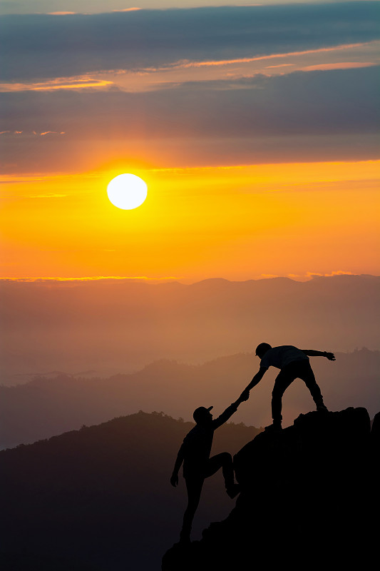 在日落时，剪影男子帮助朋友爬上岩石图片素材