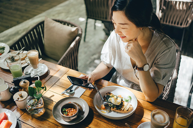 微笑的年轻亚洲妇女使用手机在一个户外餐厅吃早午餐对美丽的阳光图片素材