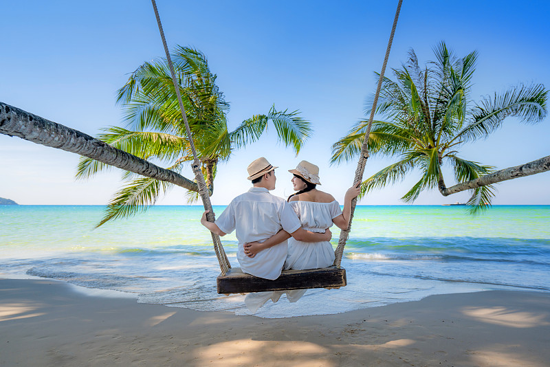 亚洲情侣情侣穿着白色礼服在海滩享受蜜月和长假，一起坐在秋千上放松和舒适，情人。夏季，旅游，假期和假日概念。图片下载