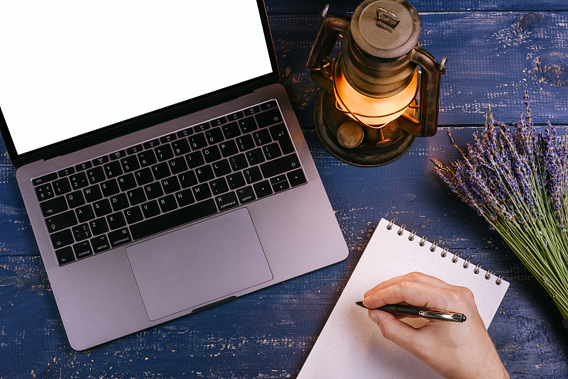 蓝色的桌子上放着一台空白屏幕的笔记本电脑，旁边放着一束薰衣草和一盏煤油灯。旁边是一个用牛皮纸写的空白笔记本。在记事本上，那人的手握着一支笔。俯视图图片素材