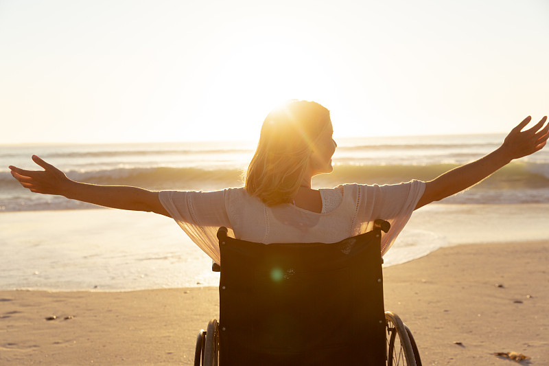 妇女在轮椅上的后视图与她的双臂在海滩上张开图片下载
