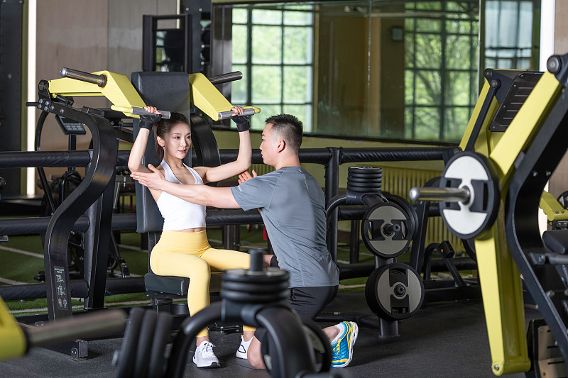 正在健身的年轻女性和男性，在健身房使用健身器材锻炼。专业健身教练指导和辅助训练、学习和交流。图片下载