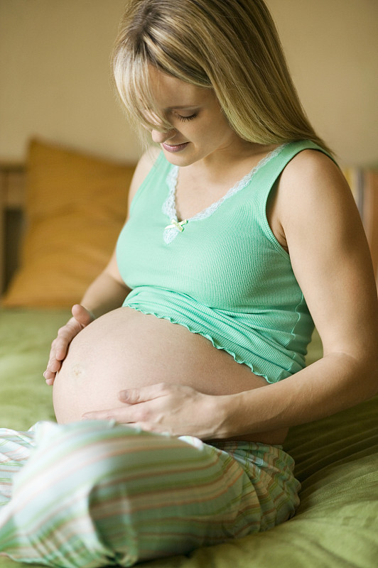 年轻孕妇坐在床上摸着肚子图片下载