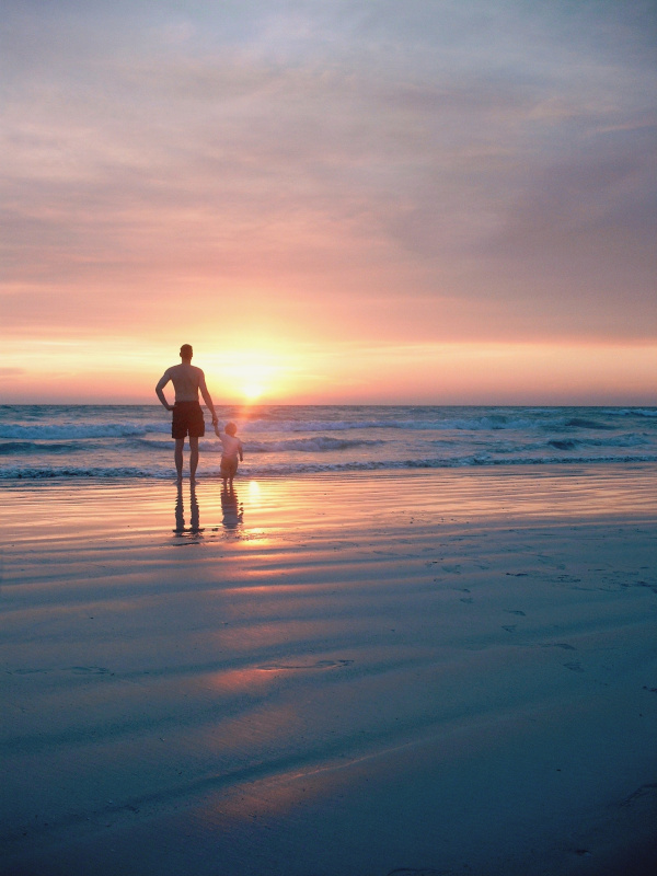 男人与孩子站在海滩上的后视图在多云的天空在日落图片素材