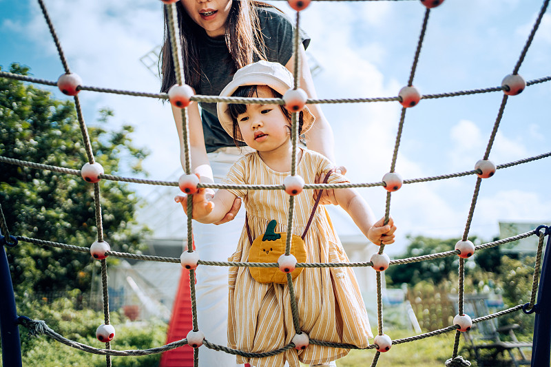 在一个阳光明媚的日子，一位年轻的亚洲母亲在户外操场上帮助小女儿攀爬绳索图片下载