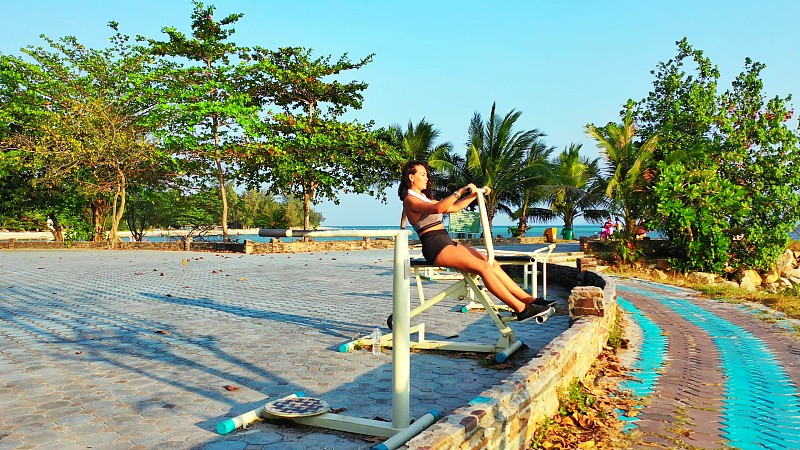 快乐的女孩在户外健身工具在美丽的度假花园周围的热带树木附近的蓝色大海图片下载