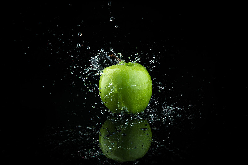 青苹果落在水中，溅起黑色背景。图片素材