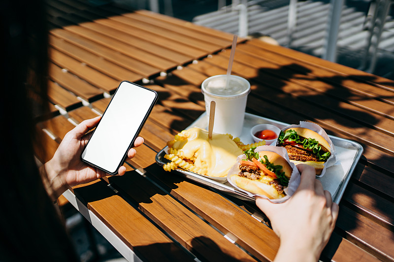 一位年轻的亚洲女性在阳光明媚的户外汉堡快餐店里用智能手机吃饭图片素材