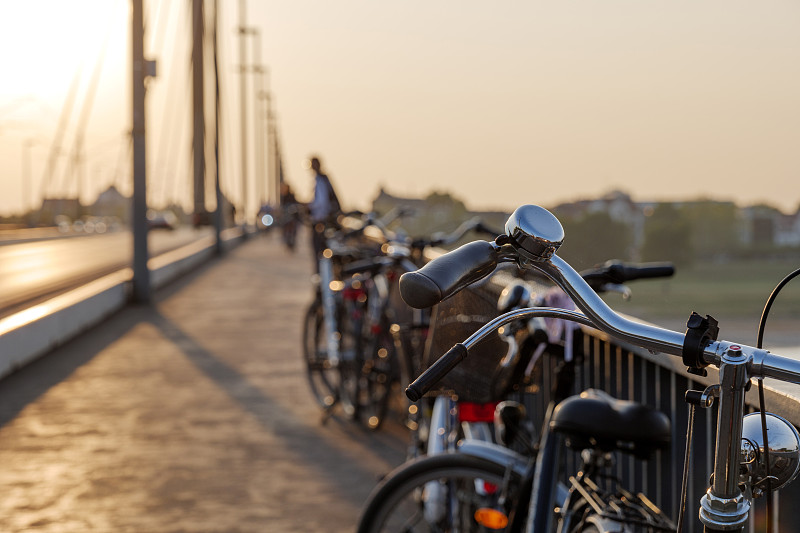在横跨莱茵河大桥的人行步道上的车把和铃铛自行车公园，以自行车、汽车、日落天空和阳光为背景的剪影低角度和选定的焦点。图片下载