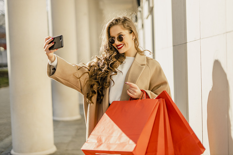 一张年轻女子在城市购物的照片，同时用她的手机自拍和微笑摄影图片下载