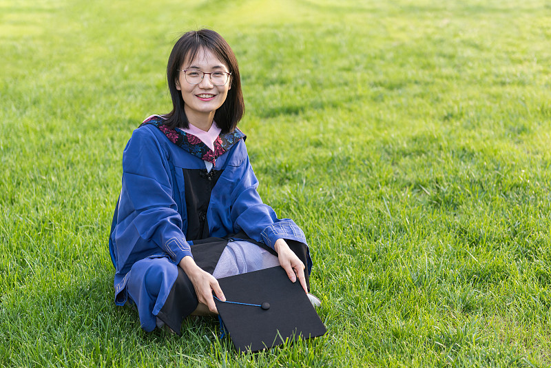 美丽的女大学毕业生坐在户外公园的绿草地上图片素材