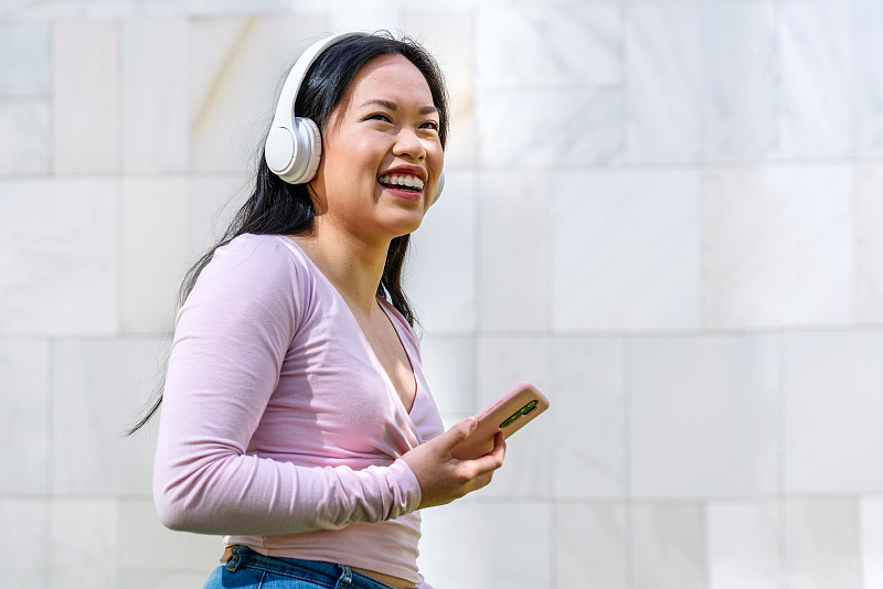 年轻快乐的亚洲女人在公园里，戴着白色耳机，一边看一边用手机图片下载