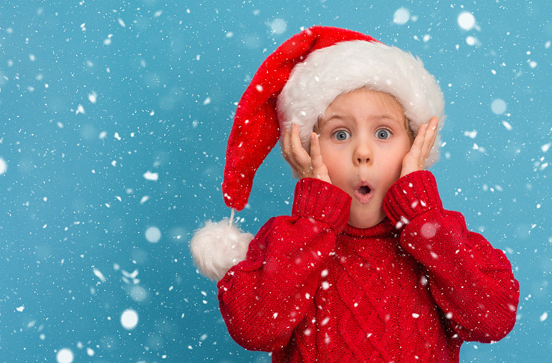 有趣的惊讶的孩子在圣诞帽和红色毛衣在蓝色的雪背景。图片下载