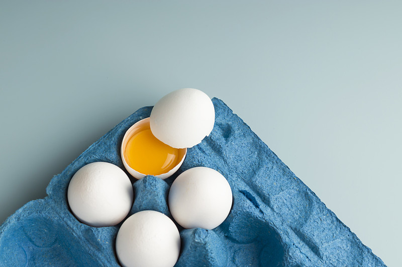 新鲜的白色鸡蛋在生态包装上的蓝色背景。蛋壳里有蛋黄的碎鸡蛋。农场的天然产品。前视图。本空间图片下载